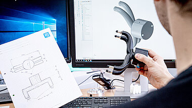 A person looks at a component and a drawing in front of a PC screen
